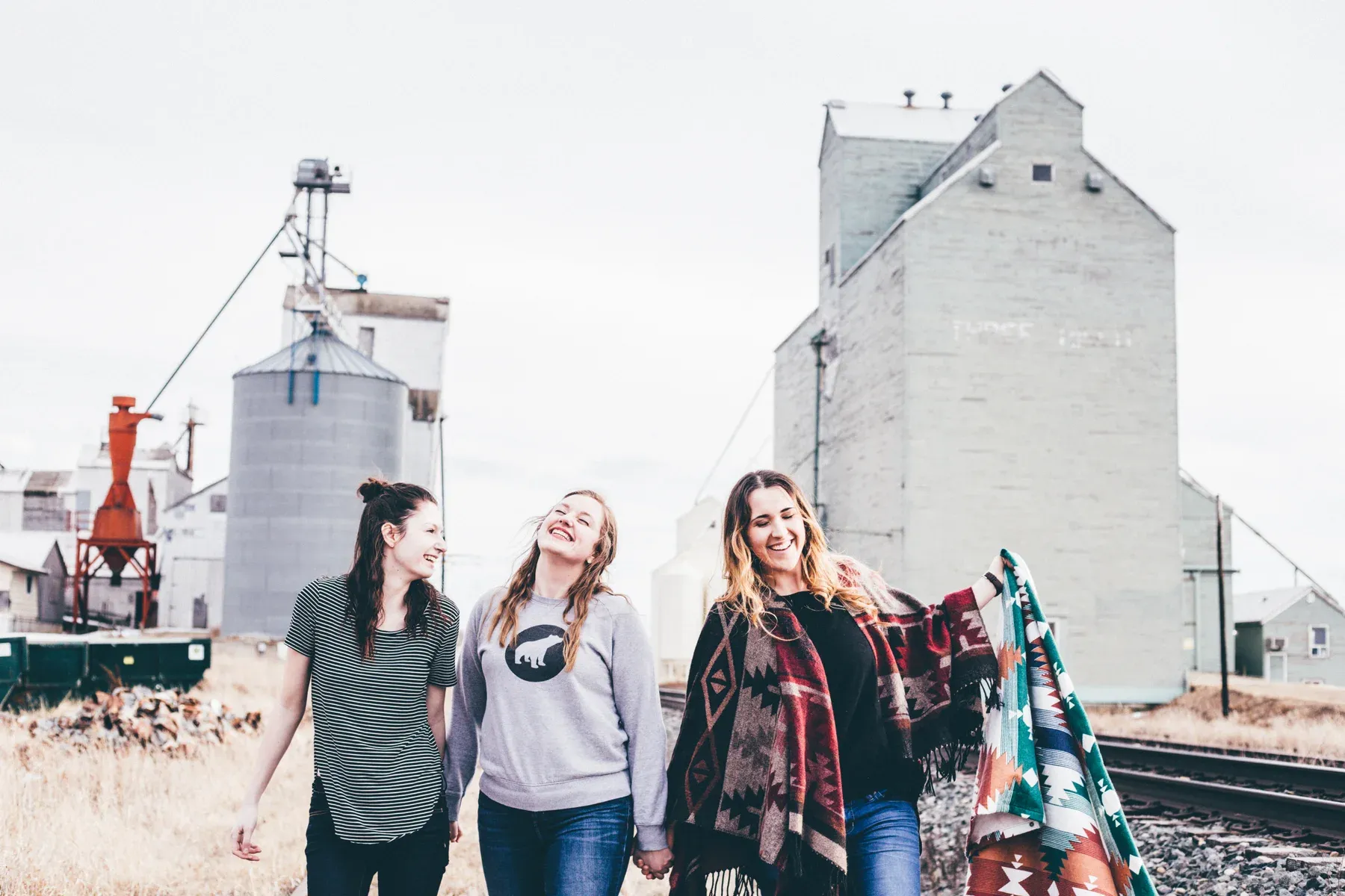 Three friends laughing outdoors, holding hands and a blanket.